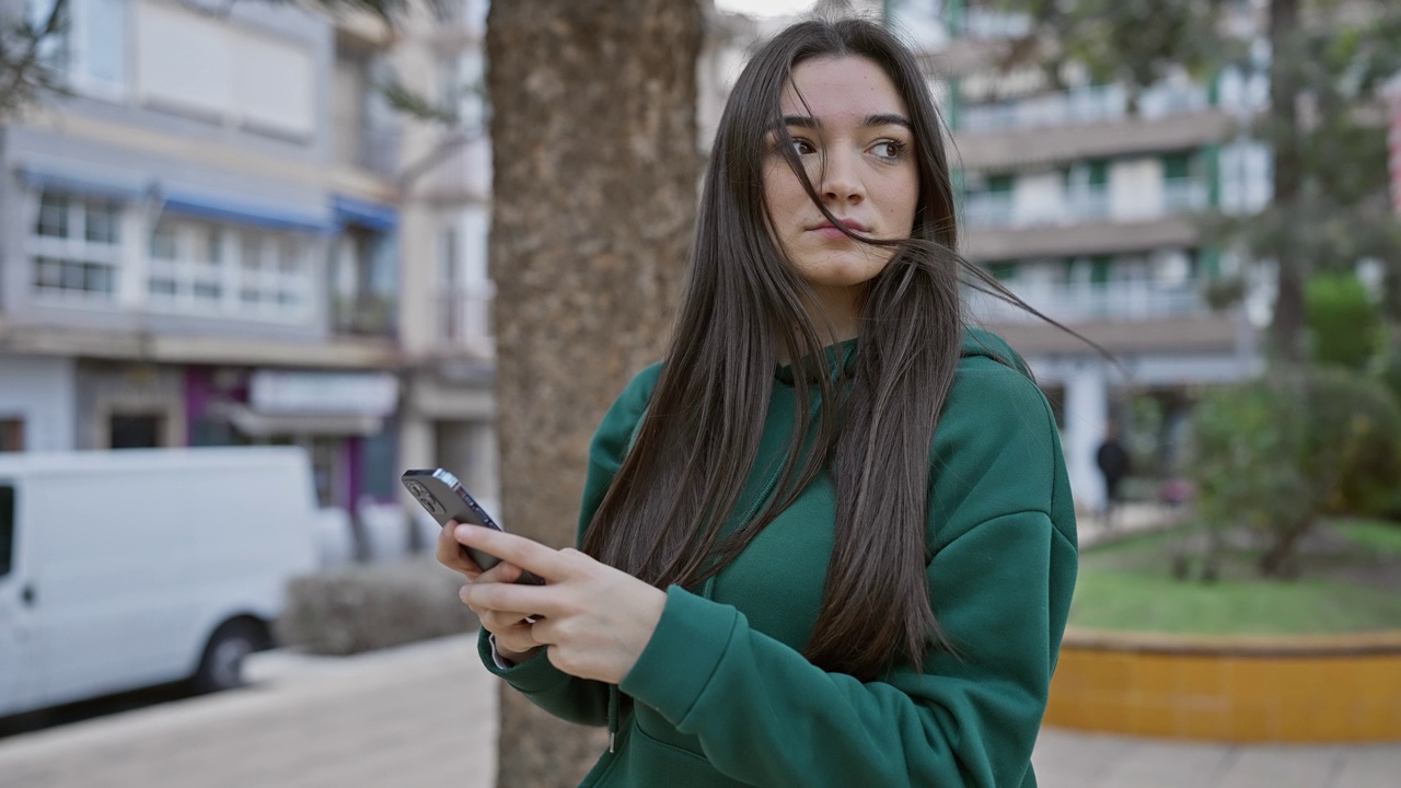 一位身穿绿色卫衣的黑发西班牙裔年轻女子在城市街道上全神贯注地玩着她的智能手机。视频素材