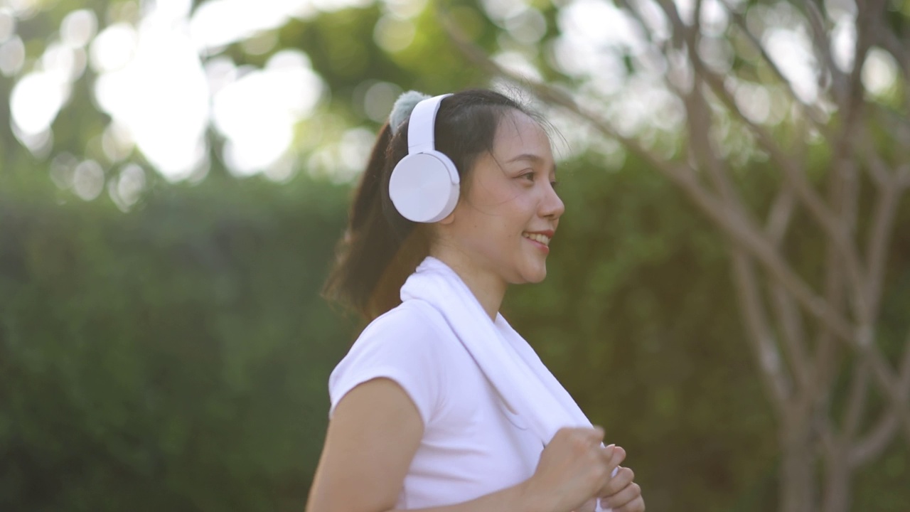 一个美丽的亚洲女人正在公园的人行道上锻炼，戴着耳机听音乐。视频素材