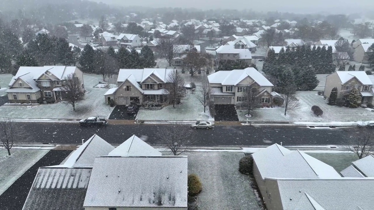 一条郊区的街道，有两层楼的房子，下着小雪，道路部分被覆盖，树木光秃秃的。航拍卡车在美国郊区附近。视频素材