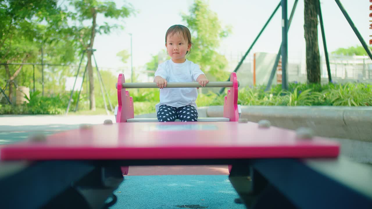 一个好奇的小孩坐在跷跷板上，手放在车把上，背景是游乐场的设备和绿色植物视频素材