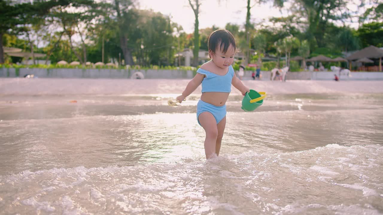 一个穿着蓝色泳衣的小孩在浅滩的水里玩着黄色的铲子和绿色的水桶视频素材