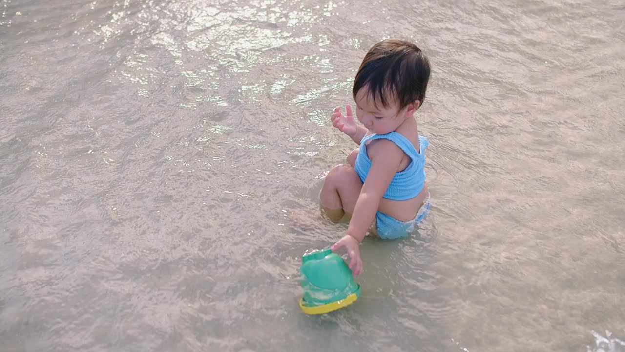 一个穿着蓝色套装的小孩在浅水里玩着一个绿色的水桶，这是一个天真的海滩乐趣时刻视频素材