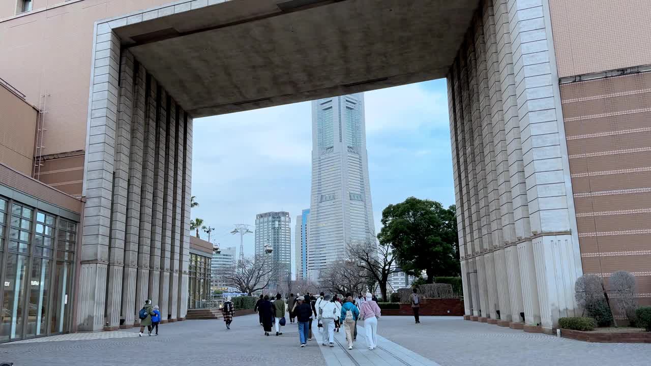 在日本横滨，人们穿过一扇巨大的混凝土大门，走向一座高耸的摩天大楼，阴天，白天的城市环境视频素材