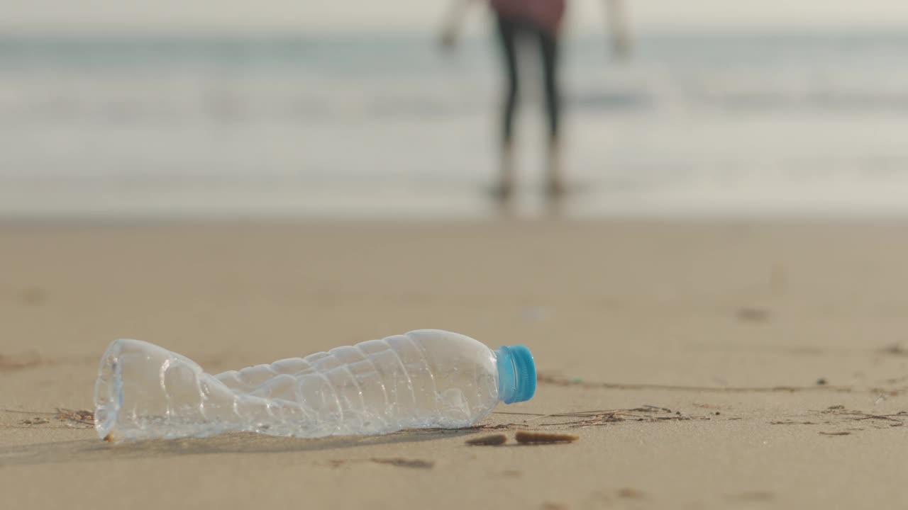 海滩上废弃的PET塑料瓶特写，以模糊的人为背景的沙滩、大海和蓝天，展示了塑料污染需要保护的生态问题概念。视频素材