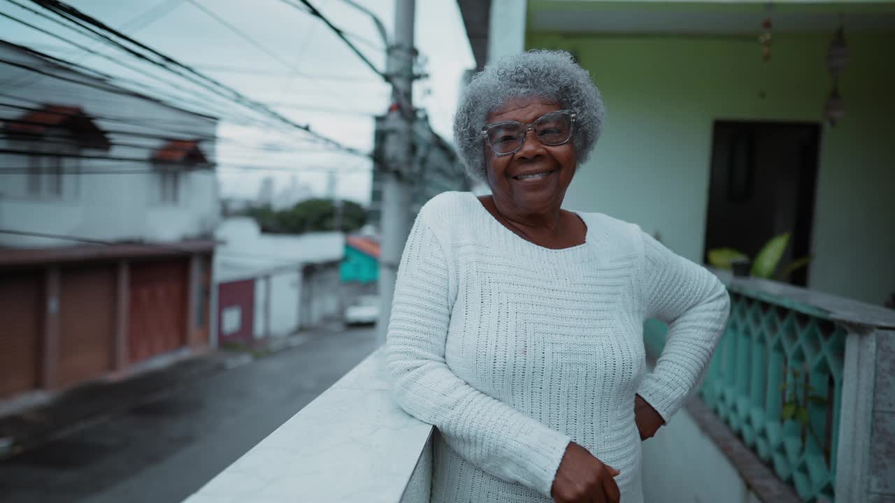 一位快乐的南美黑人老妇站在简陋的住宅阳台上，俯瞰城市街道。80年代非洲裔白发老人的肖像视频素材