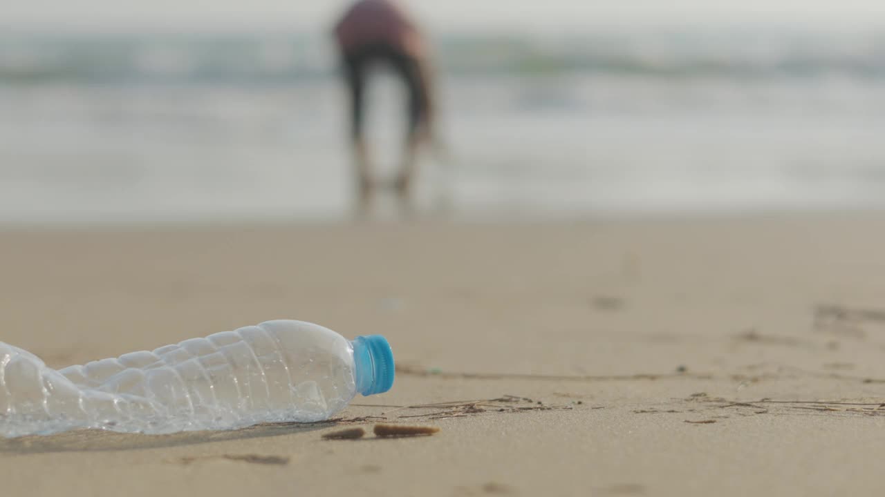 海滩上废弃的PET塑料瓶特写，以模糊的人为背景的沙滩、大海和蓝天，展示了塑料污染需要保护的生态问题概念。视频素材