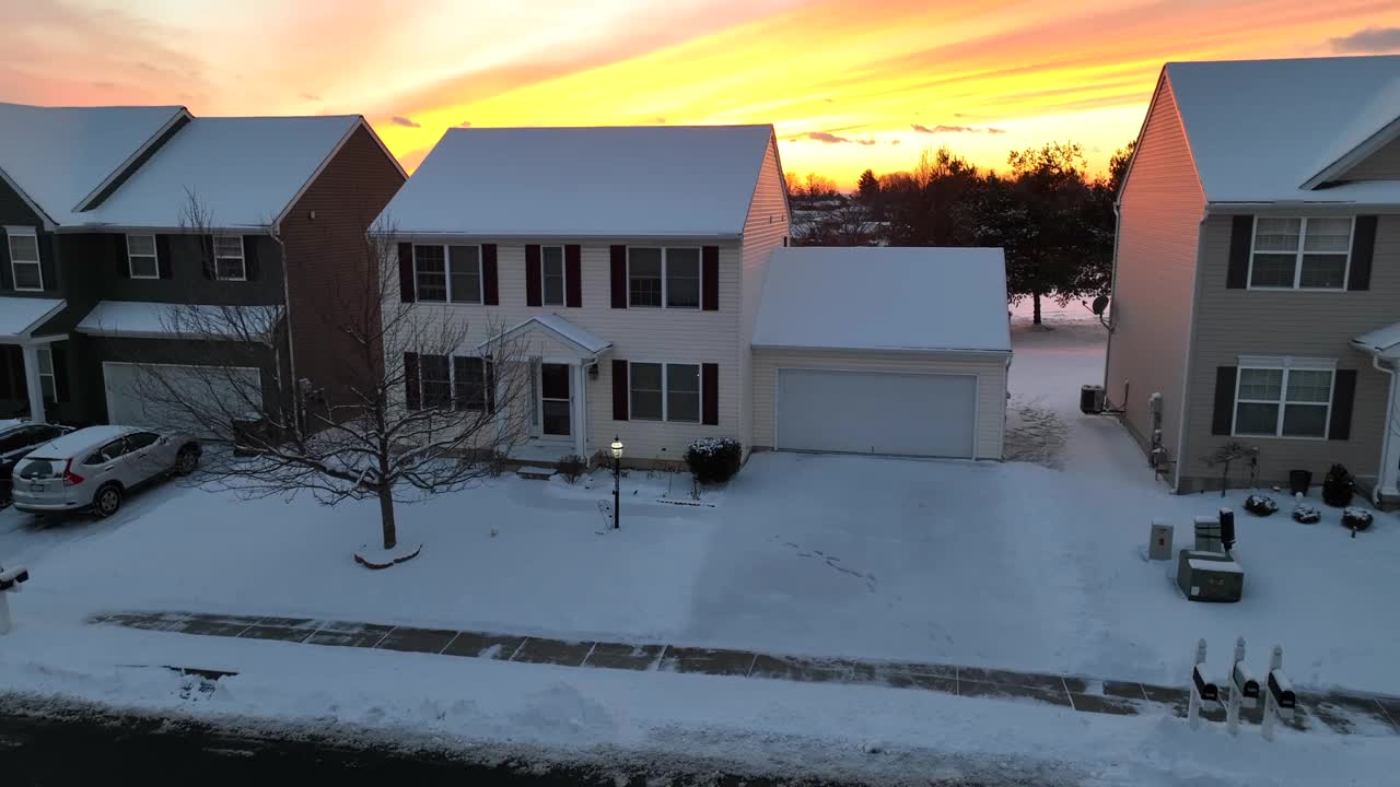 美国的邻居在冬天橙色的夕阳下被雪覆盖。单户两层住宅的空中上升镜头，揭示了美丽的天空和雪景。视频素材