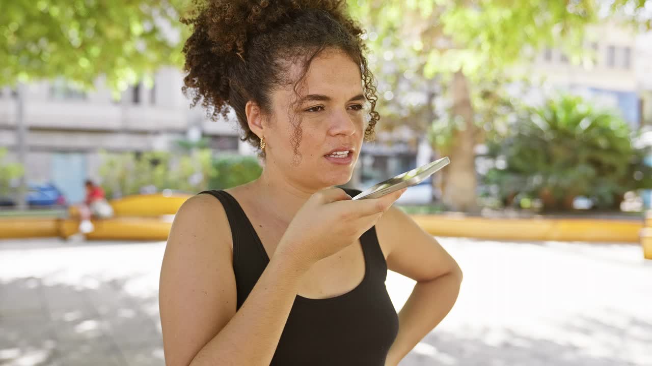 在一个阳光明媚的户外公园里，一位卷发的年轻西班牙女子正在用手机聊天。视频素材