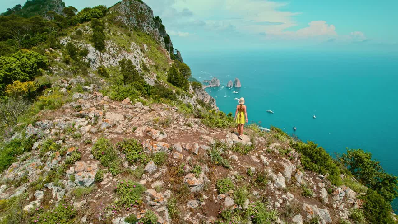 在卡普里岛山顶欣赏海景的女人。绿意盎然的岩石峭壁，映衬着清澈的海水和下方海湾中的休闲船只。夏天意大利。视频素材