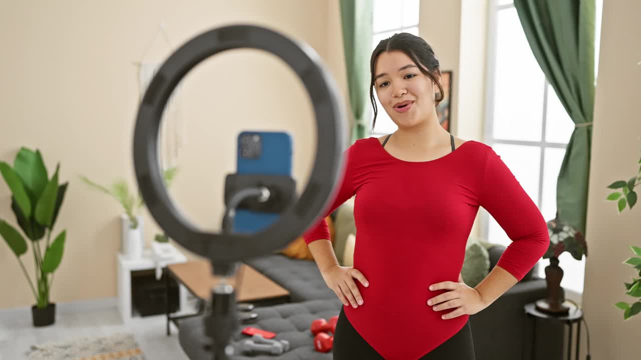 一名穿着红色衣服、面带微笑的年轻女子在家里录制健身视频，背景是一盏环灯，显示出积极的一面。视频素材