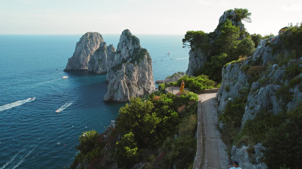 一名女子在一条宁静的小路上徒步旅行，俯瞰着宁静的地中海水域中带有法拉格里奥尼岩石的卡普里岛。意大利的夏日海景。视频素材