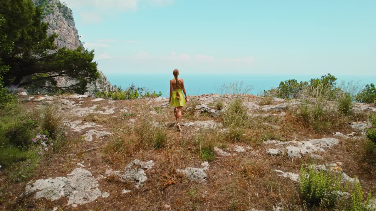 一个穿着裙子的女人在卡普里岛的悬崖上徒步旅行，凝视着第勒尼安海的壮丽景色。雄伟的法拉格里奥尼三层岩层。视频素材