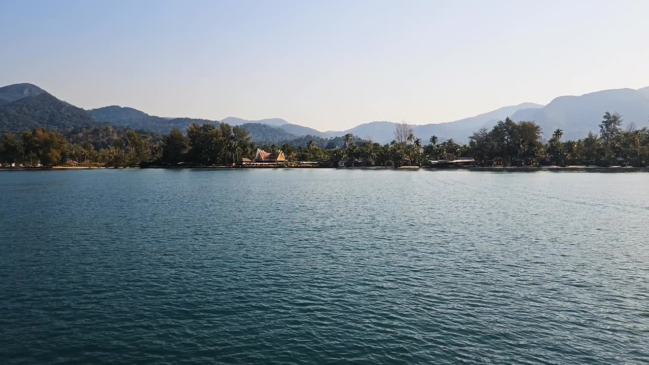 阳光明媚的热带海岸线，柔和的海浪和晴朗的天空，由郁郁葱葱的绿色植物和山脉构成视频素材