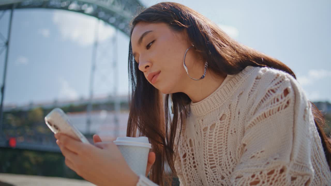 放松的女人冲浪电话在阳光明媚的河岸特写。喝咖啡的女孩视频素材