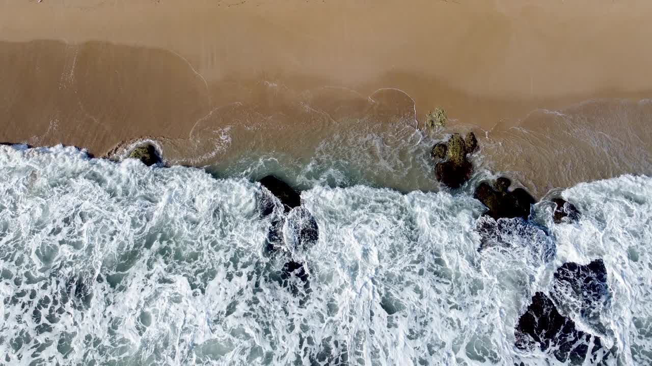 汹涌的海浪与沙质海岸相遇，捕捉水陆动态界面，鸟瞰图视频素材