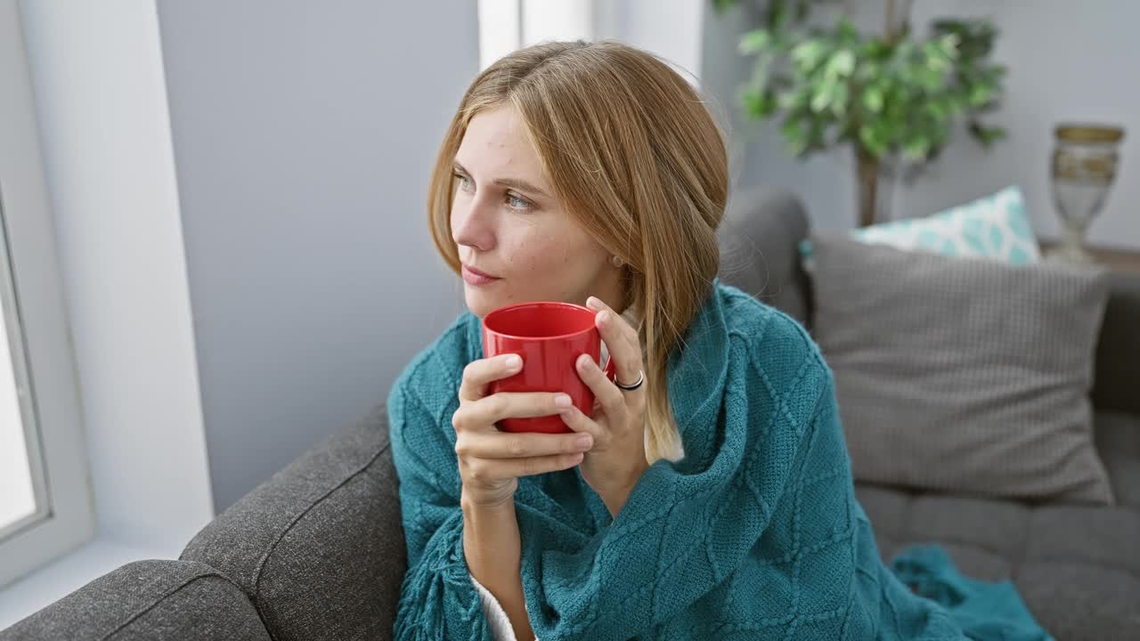 一个宁静的女人在舒适的客厅里享受一杯热饮，散发着舒适和放松的气息。视频素材