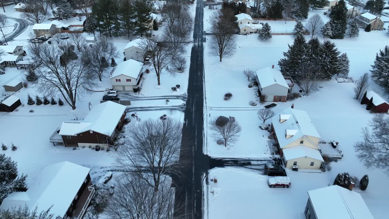 航拍到的白雪皑皑的街区，道路两侧都是树木。无人机向上倾斜，露出美国房屋上空的冬日黄昏。视频素材