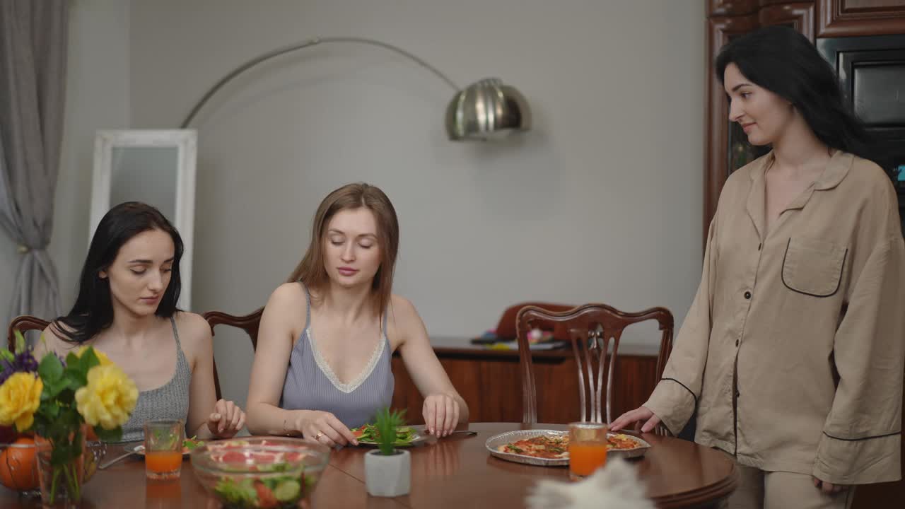 漂亮的年轻女子开心地吃着披萨，把新鲜的蔬菜沙拉放在一边，坐在客厅的桌子旁。女孩们微笑着，享受着手中披萨的味道。视频素材