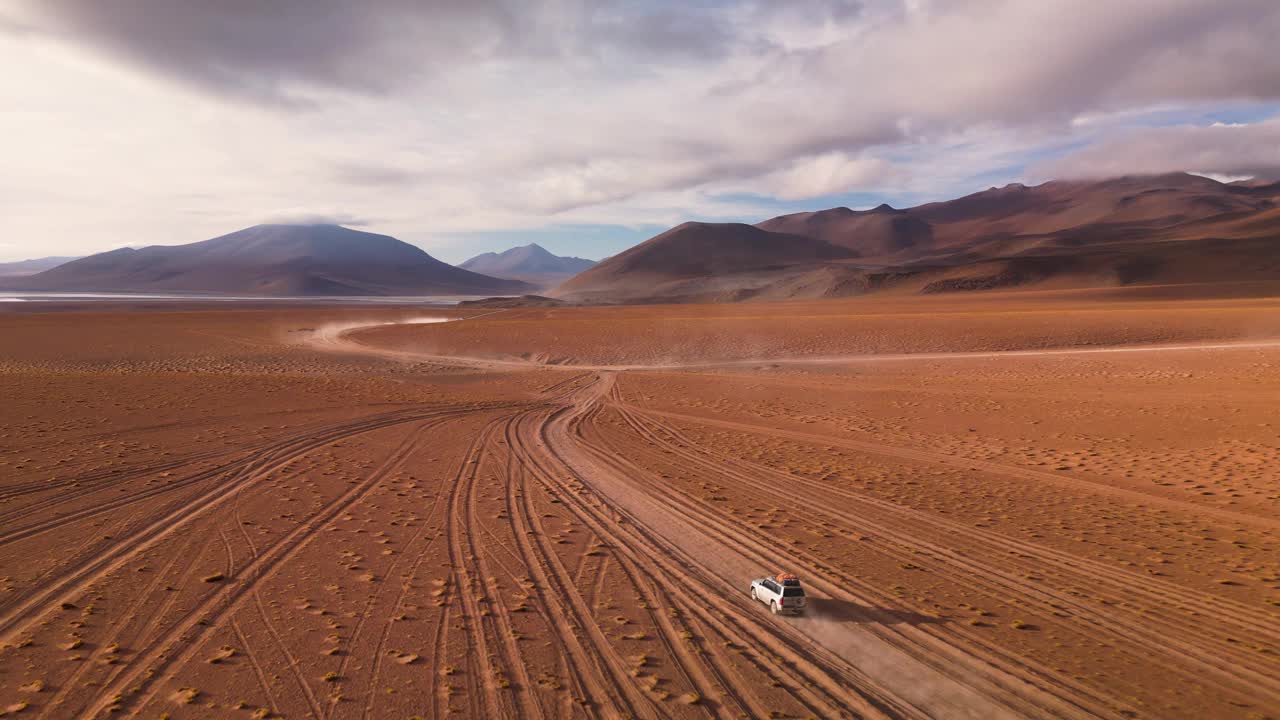 白色SUV行驶在尘土飞扬的道路上，广阔的沙漠和山脉在晴朗的天空下，鸟瞰图视频素材