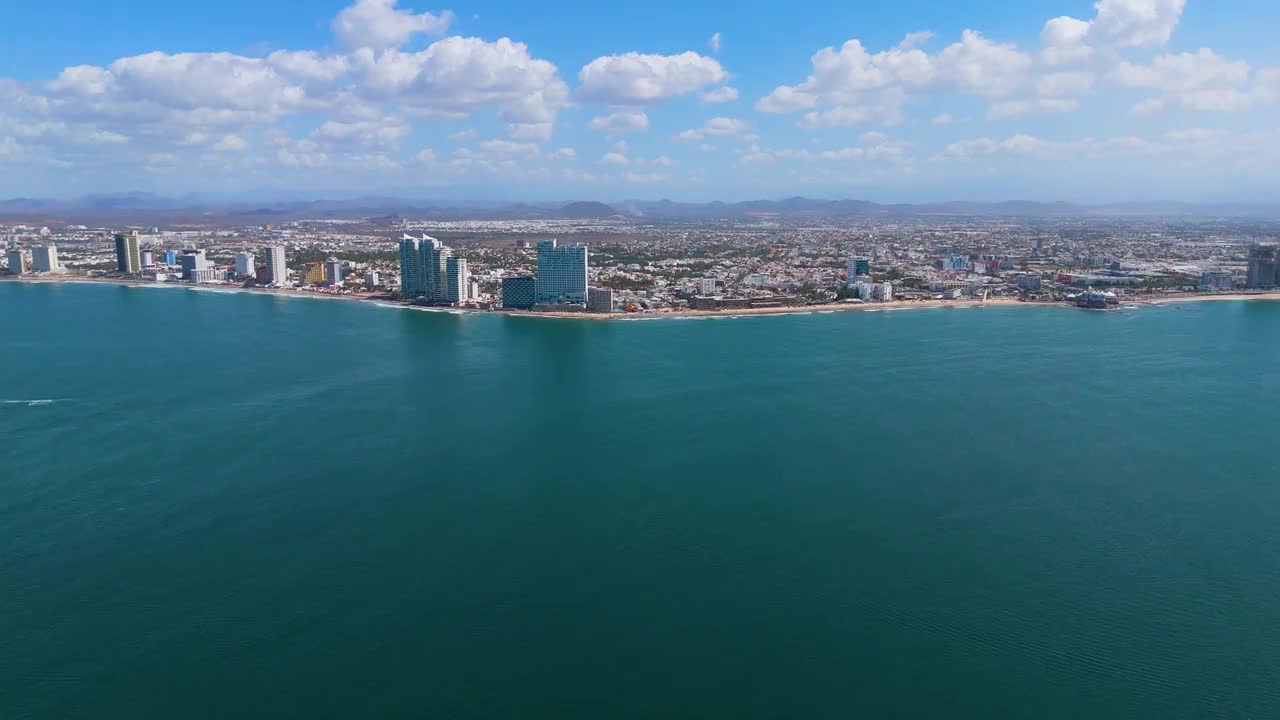 从荒岛到墨西哥马萨特兰海滩的惊险空中之旅。游船在蓝色的海面上留下白色的尾迹，增强了壮观的景色。当无人机靠近海滩时，突出的酒店增加了这个墨西哥人的宏伟视频素材