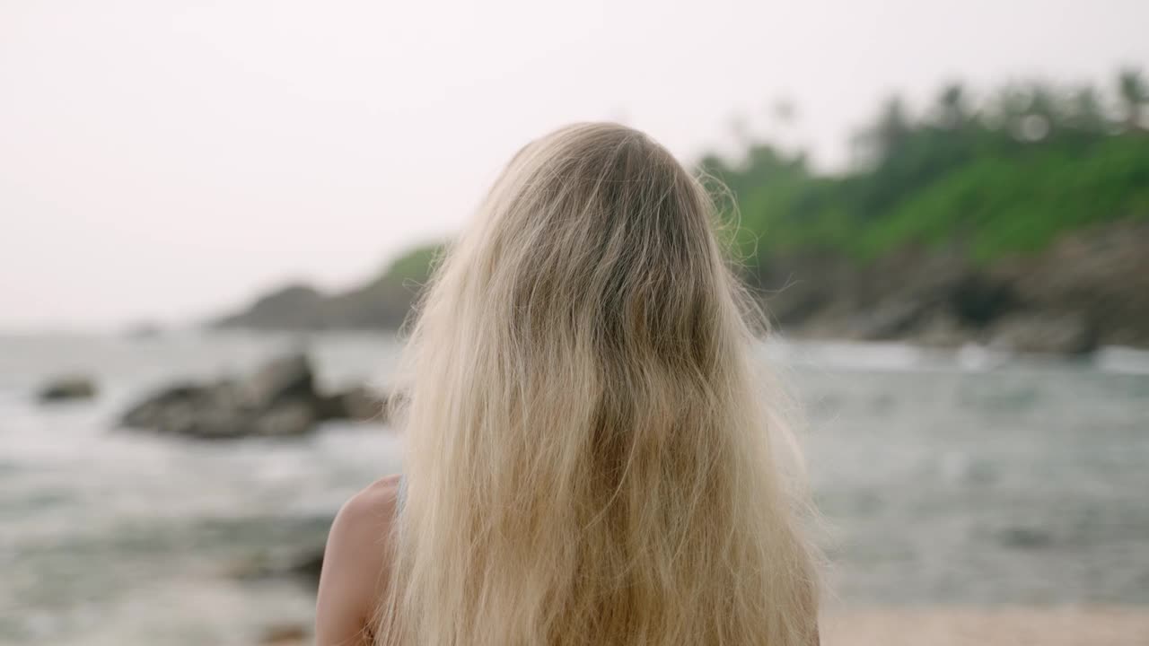 金发女郎走在热带海滩上，海浪拍打着海岸线。身着夏装的女性在海岛海岸享受大自然。浪漫度假，休闲散步，自然海景背景与岩石。Slowmo视频素材