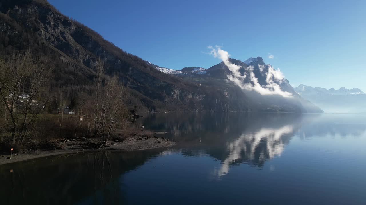 在晴朗的日子里，无人机朝着雾蒙蒙的雪顶山脉，在平静的湖边移动视频素材