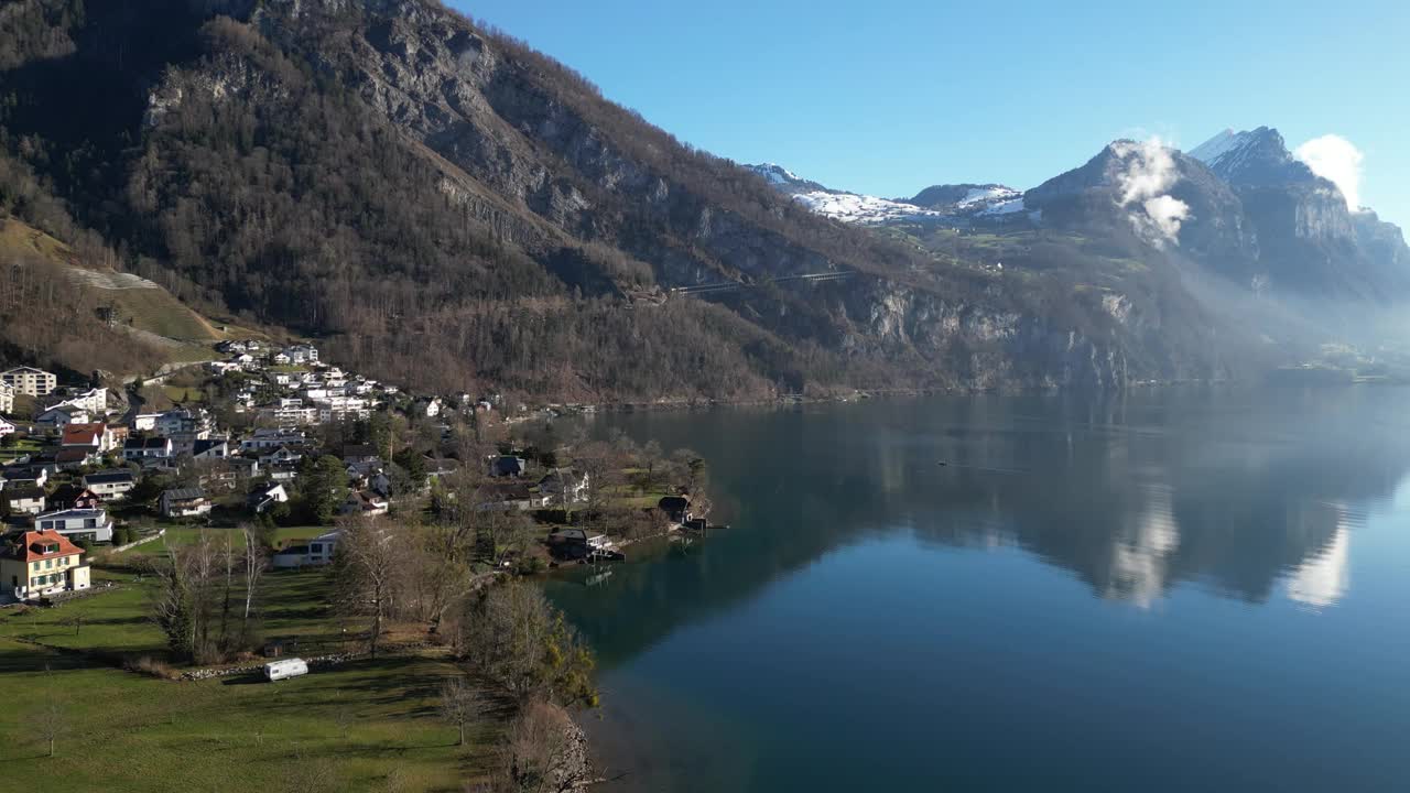 宁静的无人机剪辑显示平静的湖泊，在海岸线上的村庄，和雪山的背景，在一个阳光明媚的春日视频素材