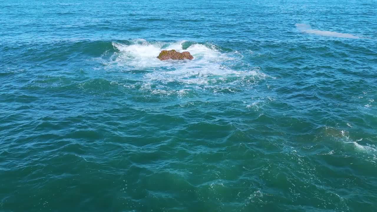 在浩瀚的海洋中，一群岩石浮出水面，被翻滚的海浪轻轻地亲吻着。当波浪抚摸着它们的表面时，缕缕白色的浪花在空中飞舞，创造了一个令人着迷的自然美景。视频素材