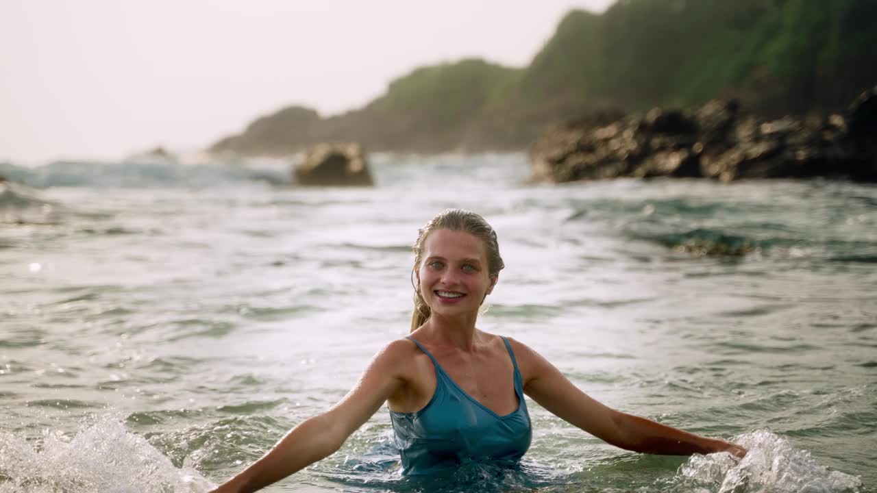 身着蓝色连衣裙的优雅女子在日落时分享受海浴。微笑的女士在温暖的海水中放松，海水在浪漫的风景海滩上飞溅。穿着飘逸，体验大自然的宁静。缓慢的运动。视频素材