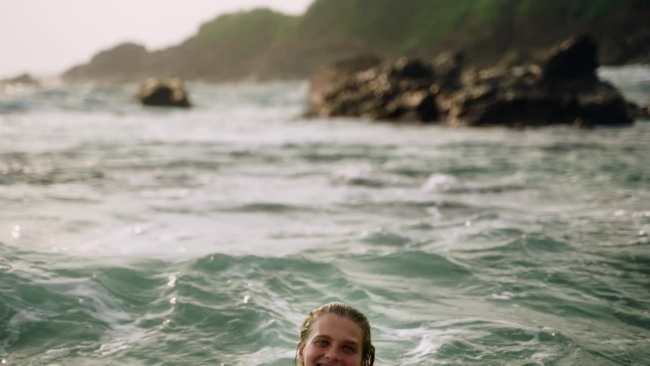 微笑的穿着裙子的女人享受温暖的海水，在阳光明媚的海滩上嬉戏飞溅的水。浪漫无忧无虑的独自旅行者享受海浪，自由。风景优美的海岸背景衬托出欢乐的时刻。Slowmo视频素材