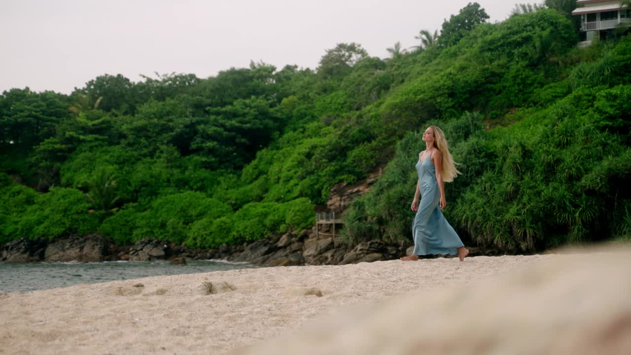 身着蓝色连衣裙的优雅女子漫步在幽静的海滩上。金发，赤脚奢华，专属隐居。海风吹来，安安静静的走，宁静的岸。独自旅行者享受宁静的海岸，自由。Slowmo视频素材