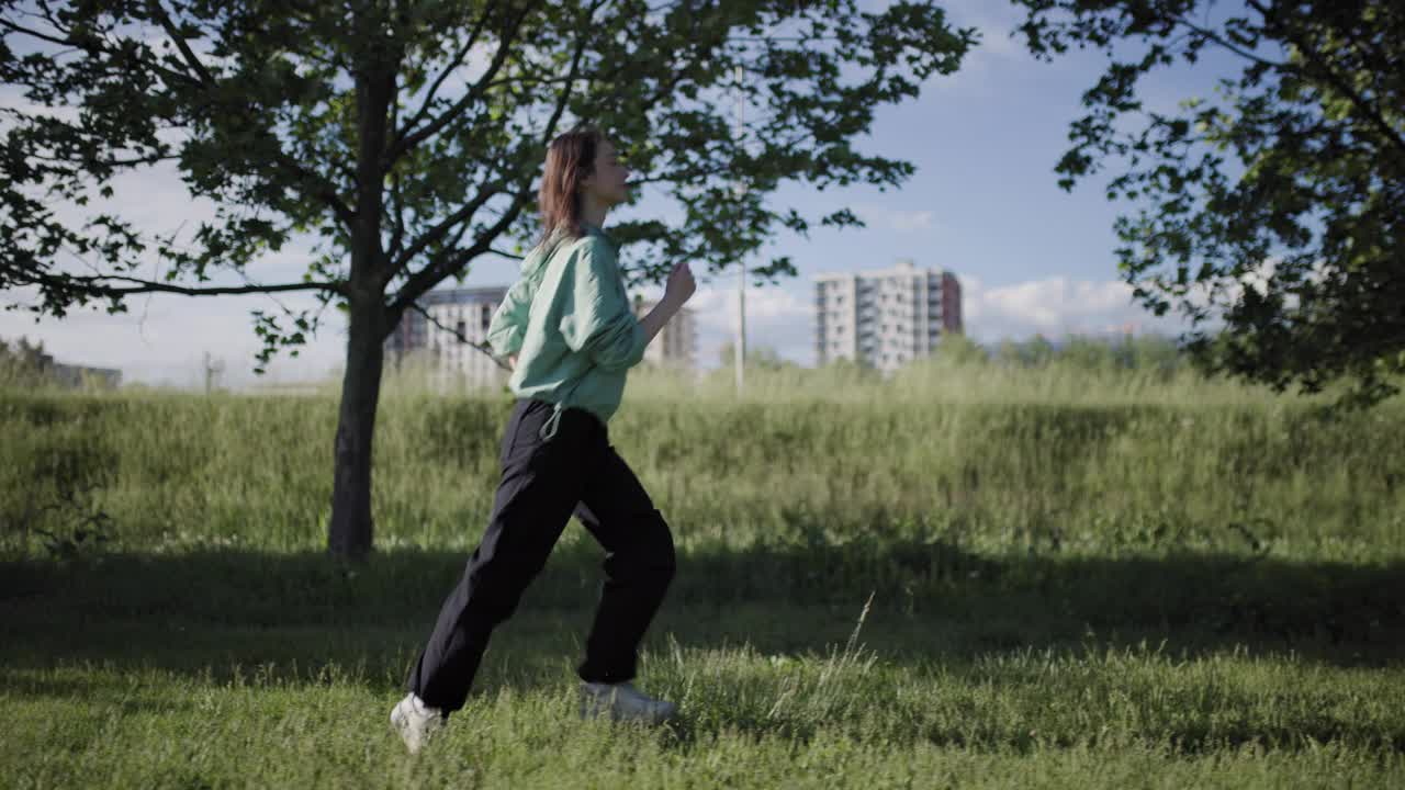 一个活跃的年轻女子沿着郁郁葱葱的公园小路奔跑，蓝天下绿树成荫，远处是城市的天际线。视频素材