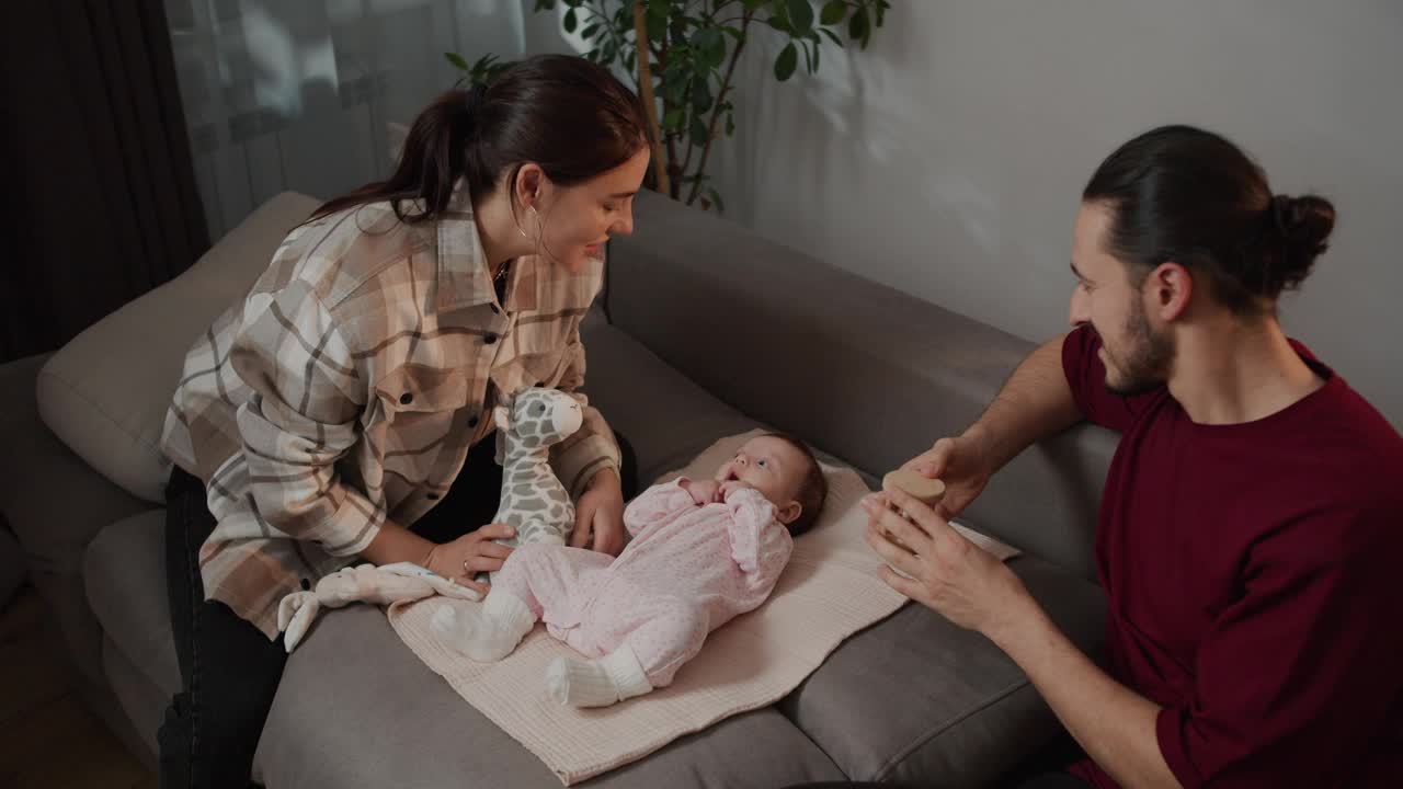 快乐的黑发女孩穿着格子衬衫和她的黑发丈夫一起穿着红色t恤和他们的小女儿玩各种各样的儿童玩具，就像一个婴儿穿着粉红色的工装裤坐在灰色的沙发上在现代公寓视频素材