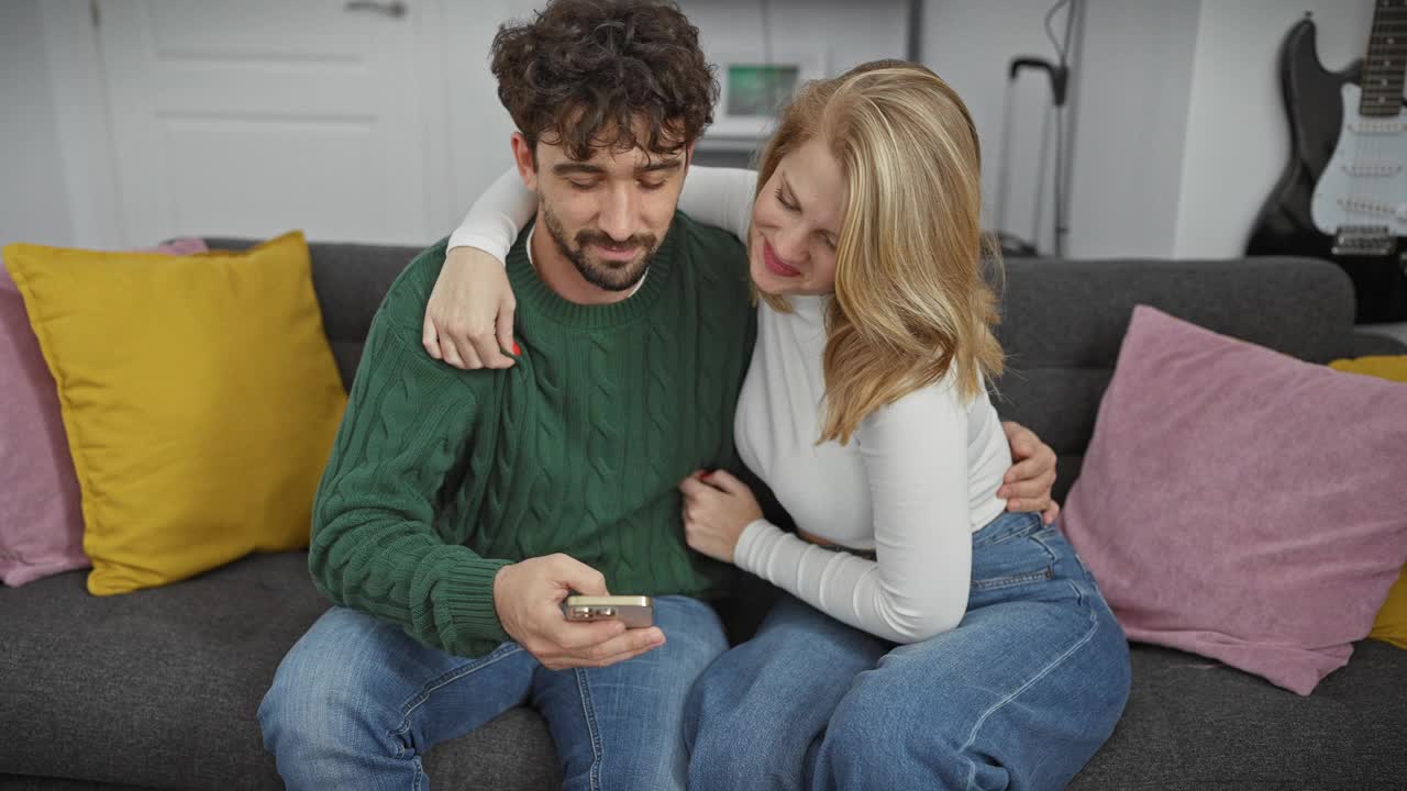 在一个舒适的客厅里，一个微笑的女人亲吻了一个拿着智能手机的男人，两人都坐在沙发上，沙发上有彩色的枕头。视频素材