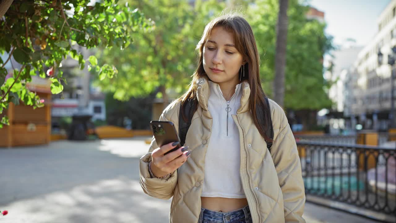 一位年轻女子在阳光明媚的城市公园里用智能手机自拍。视频素材