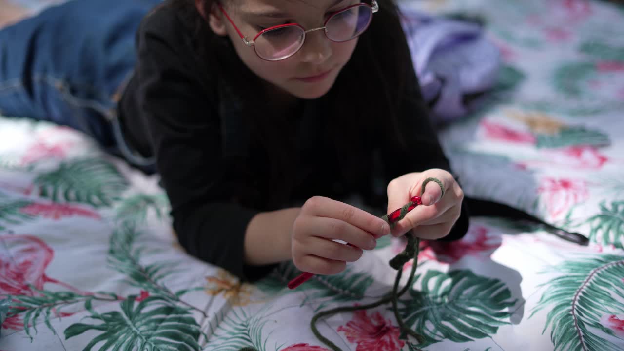 一个戴着眼镜的漂亮小女孩躺在公园野餐的毯子上，用编织针和线编织。户外爱好视频下载