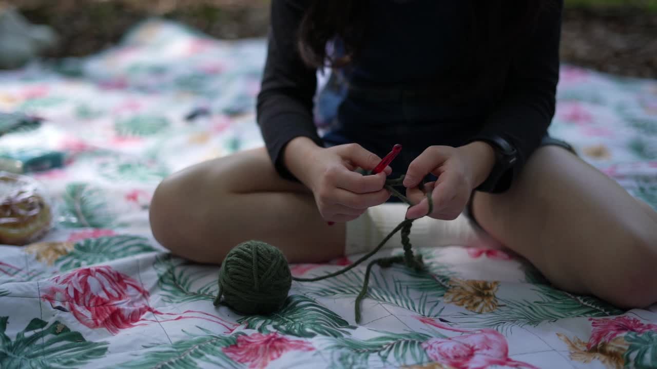 一个戴着眼镜的漂亮小女孩坐在公园里野餐，像瑜伽士一样翘着二郎腿，用编织针和线编织。户外爱好视频下载