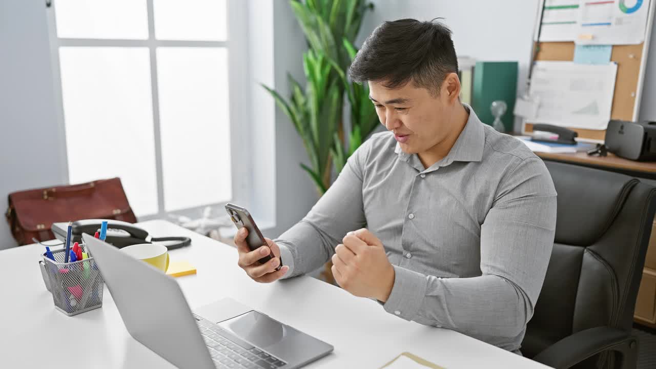 年轻的亚洲人在办公室用智能手机，笔记本电脑放在桌子上。视频素材