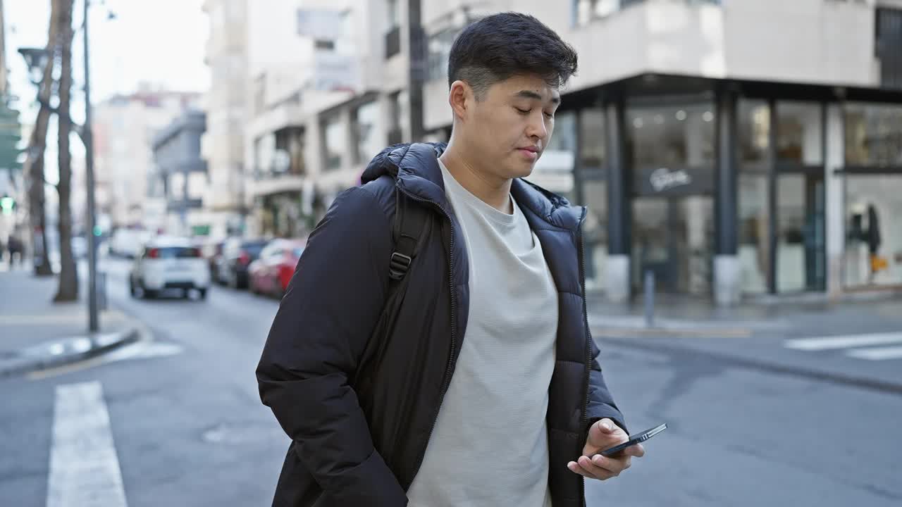 一个穿着夹克的年轻亚洲男子在熙熙攘攘的城市街道上使用电话。视频素材