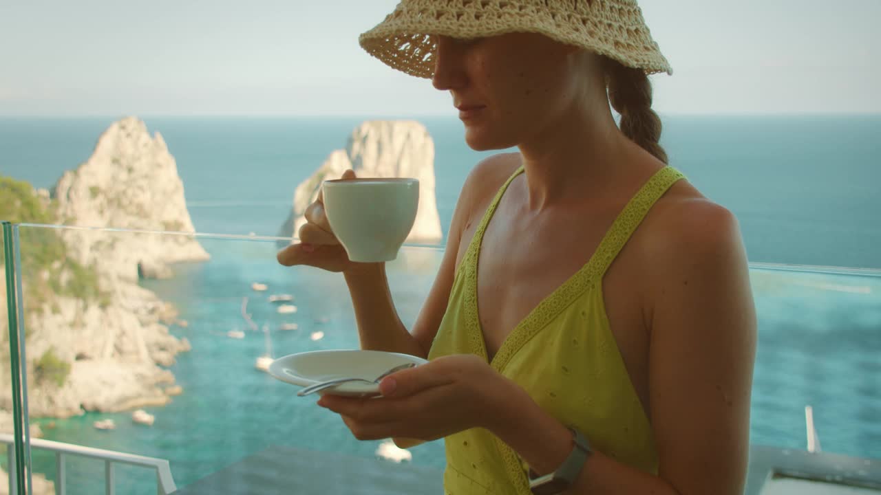 在意大利卡普里岛的夏日旅游中，一名女子在俯瞰大海和法拉格里奥尼悬崖的阳台上享受着一杯咖啡。视频素材