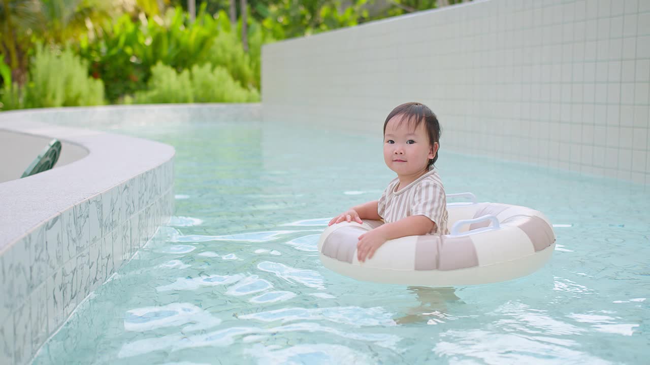 可爱的幼儿女孩戴着太阳镜漂浮在游泳池与充气环在一个现代化的度假胜地周围郁郁葱葱的绿色植物，夏季娱乐和休闲，快乐的童年时刻，户外活动视频素材