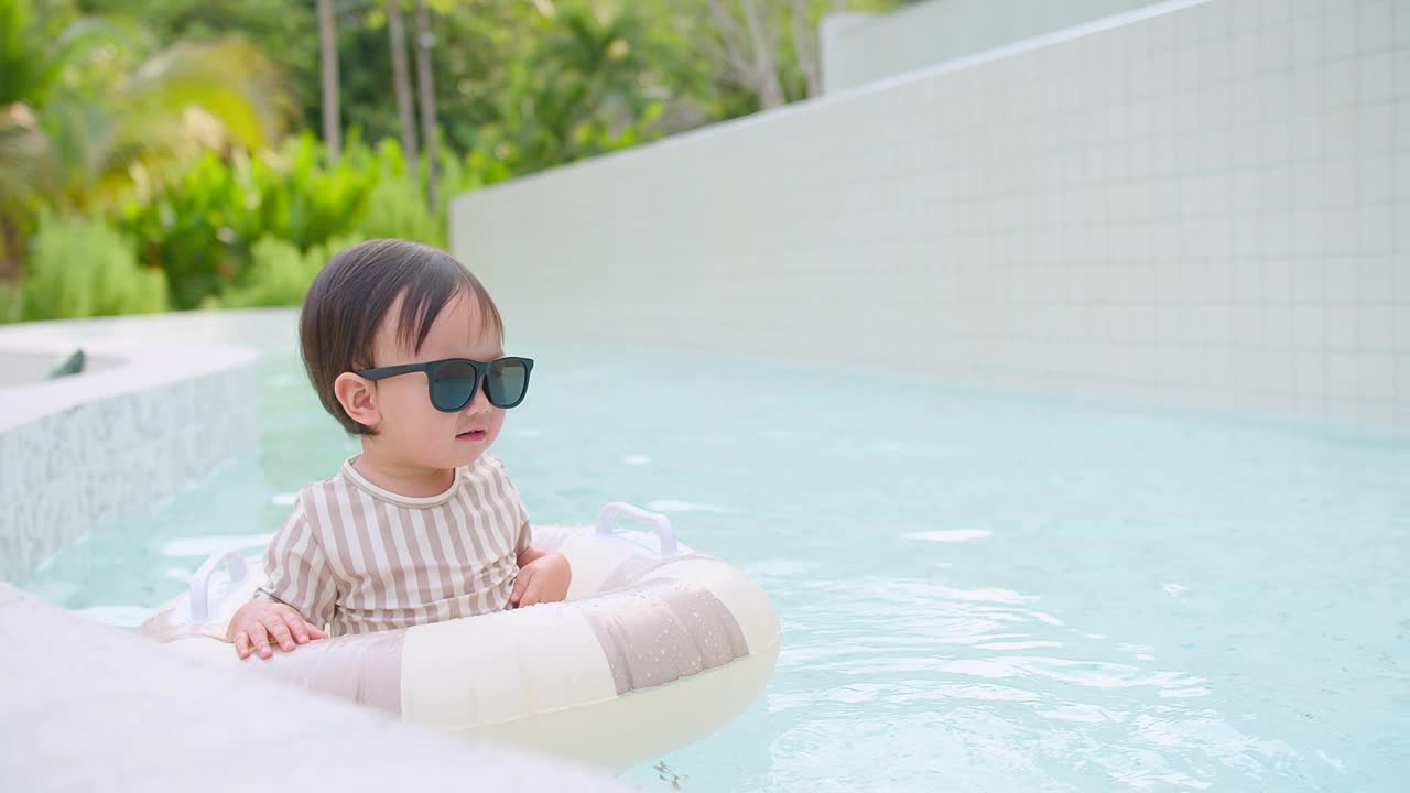 可爱的幼儿女孩戴着太阳镜漂浮在游泳池与充气环在一个现代化的度假胜地周围郁郁葱葱的绿色植物，夏季娱乐和休闲，快乐的童年时刻，户外活动视频素材