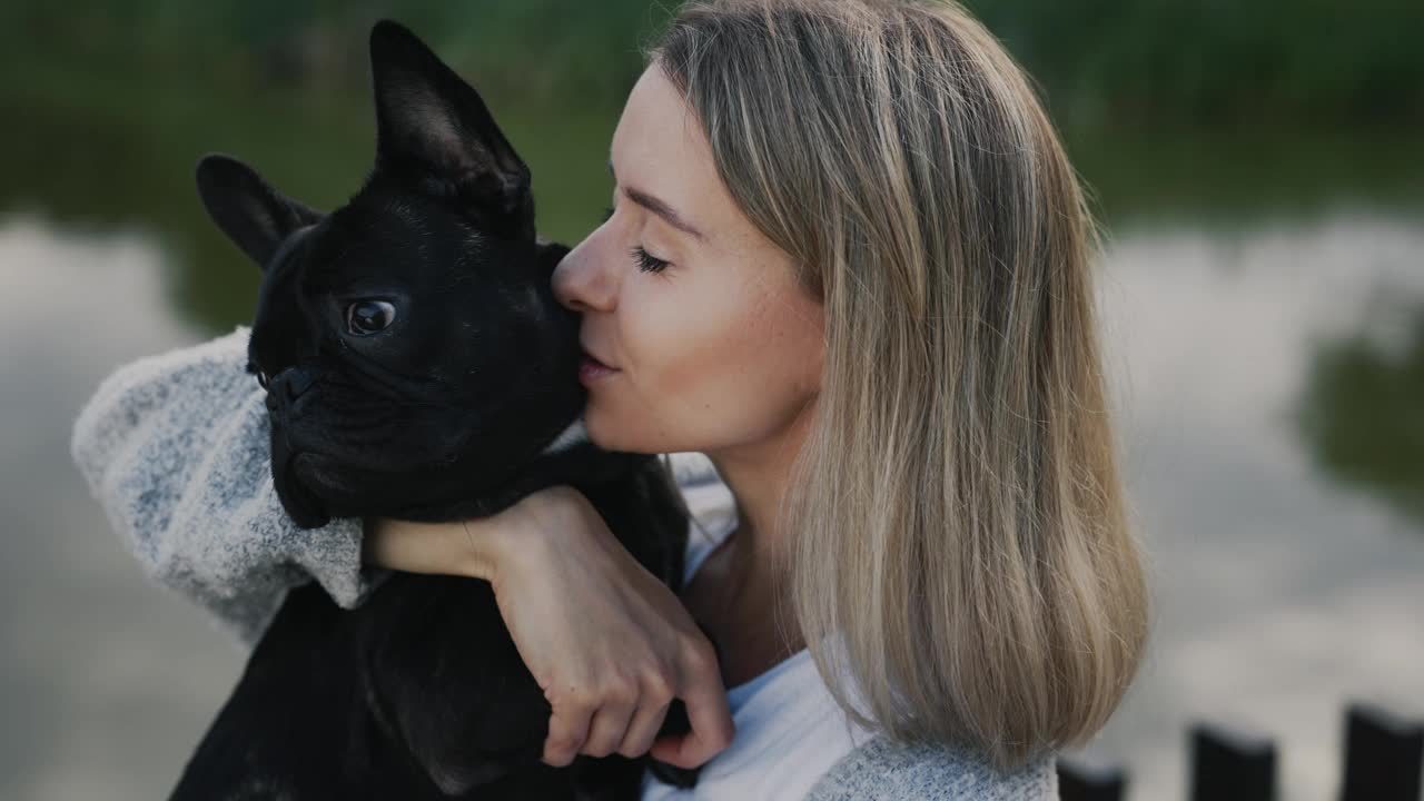 一位快乐的白人女宠物主人正在抚摸和亲吻她的黑色法国斗牛犬小狗，向站在当地湖泊附近的公园里的狗狗表达爱意。关闭了。人与狗的友谊概念。视频素材