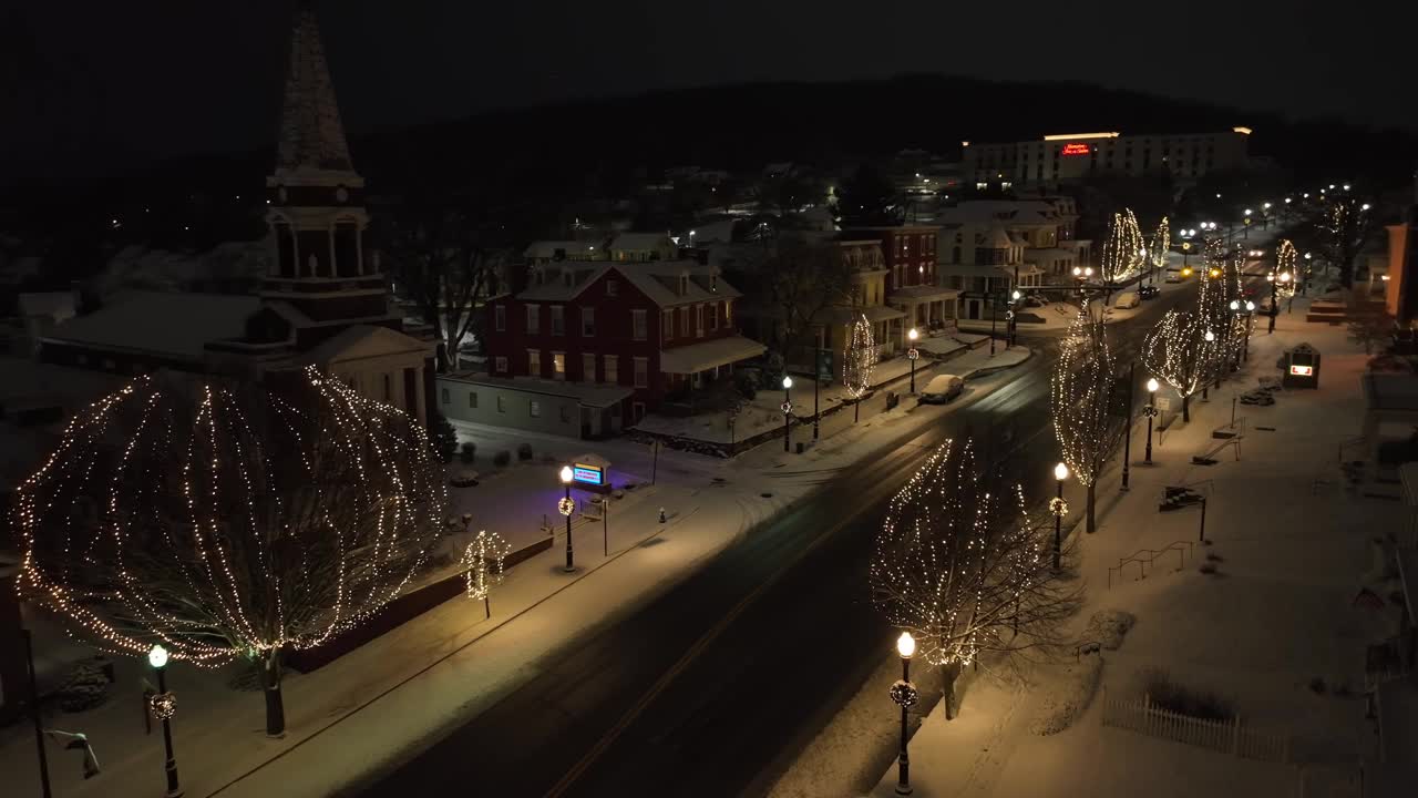 古雅的美国小镇在冬夜被雪覆盖。航拍的主要街道，房屋，教堂和圣诞树的圣诞灯。视频素材