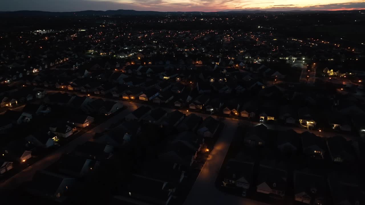 夜景鸟瞰郊区，街灯和房屋灯照亮，背景是远处的城市灯光和五彩缤纷的日落。在宁静祥和的环境中拍摄的。美国。视频素材