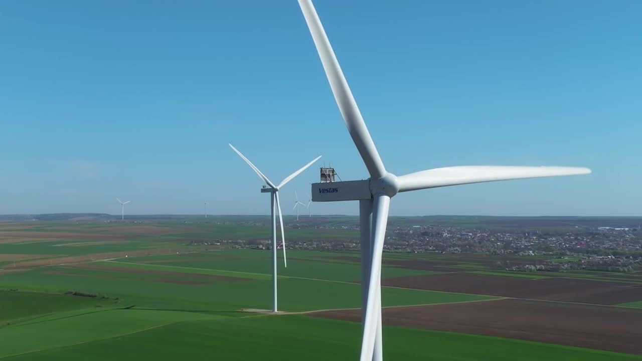 在一个阳光明媚的日子里，风力涡轮机在广阔的绿色田野和遥远的村庄上空旋转视频下载