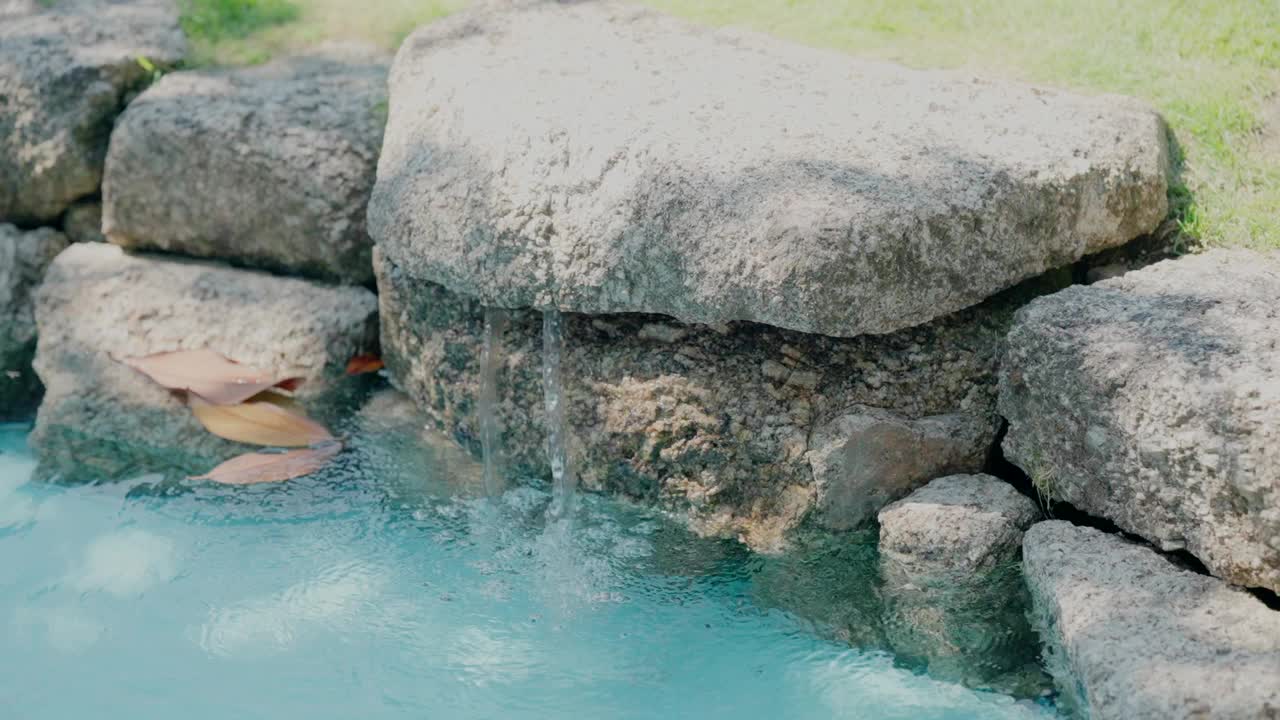 小水景，清澈的蓝色水轻轻地流过大石头，周围是郁郁葱葱的花园视频素材