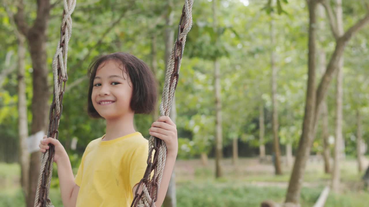 可爱的亚洲小女孩在树木环绕的户外欢快地荡秋千。它表现了童年玩耍、快乐和冒险的本质，突出了自然的无忧无虑的精神和宁静。视频素材