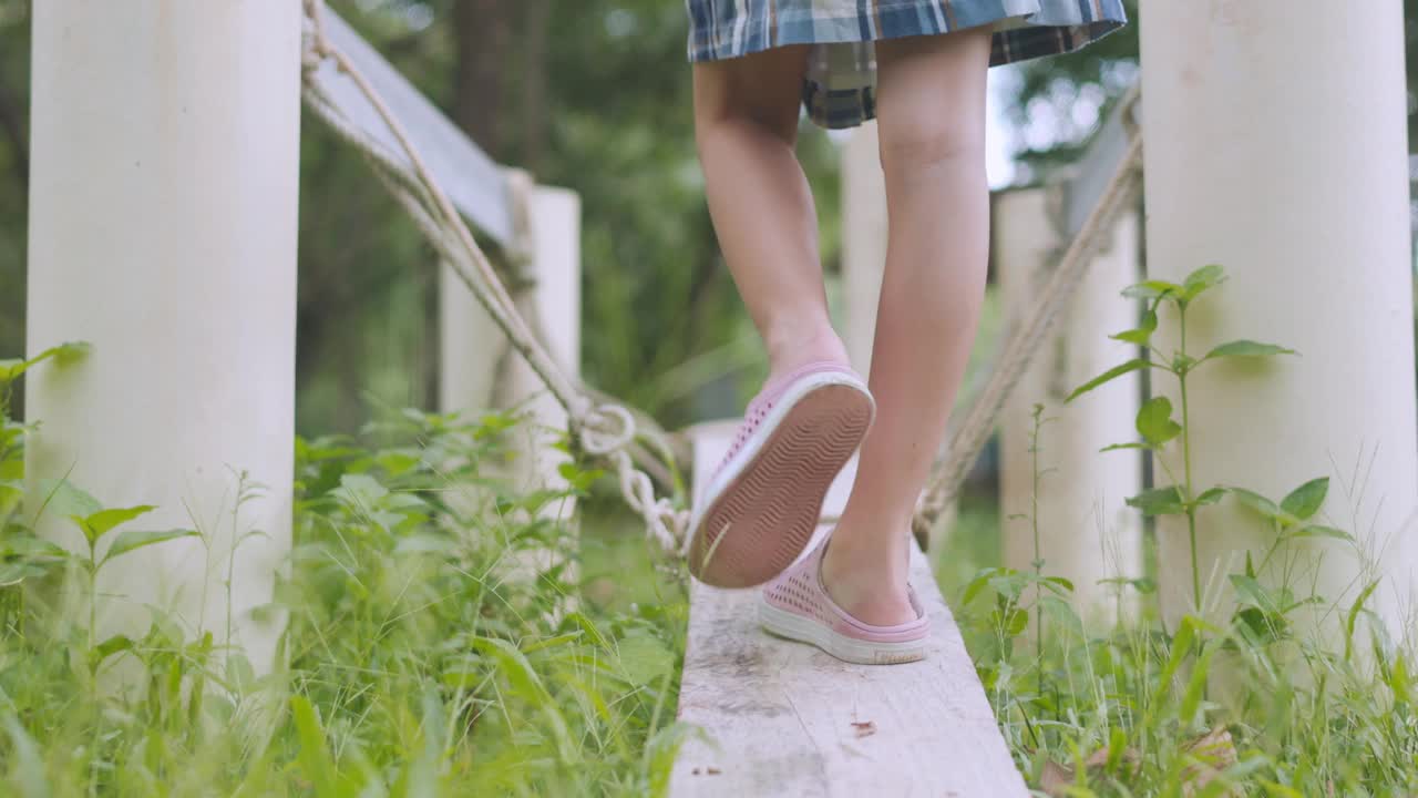 近距离，一个穿着粉色鞋子的女孩的脚站在绳桥上的低视角，象征着童年的冒险和不确定性，表现出与绿色植物一起玩耍的乐趣、探索和快乐。视频素材