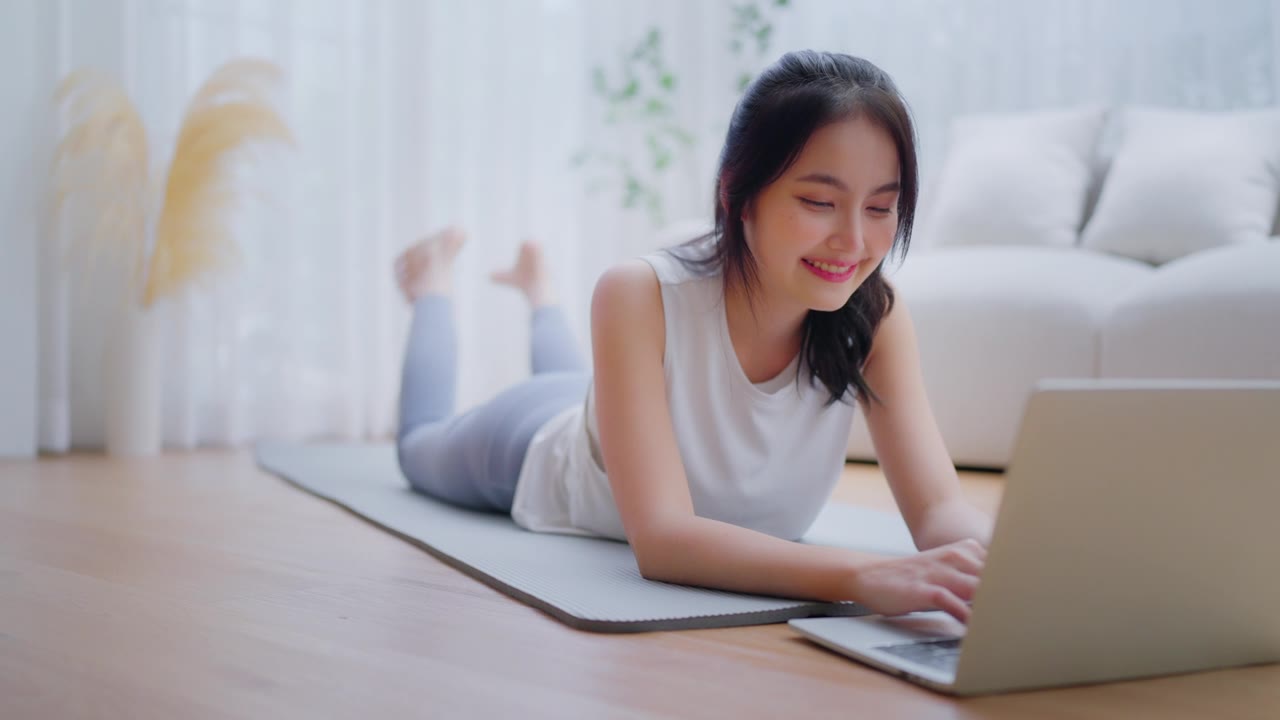 年轻的亚洲女性躺在地板上的瑜伽垫上，坐在笔记本电脑前，在家训练。美丽的微笑的女人在做伸展运动，同时在锻炼前与教练在线交流视频素材画面1